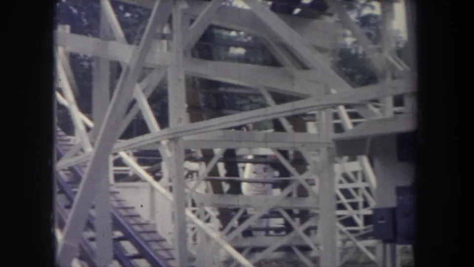 1968 Michigan amusement park frame with white wooden coaster supports and a red train carrying riders partly obscured by beams.