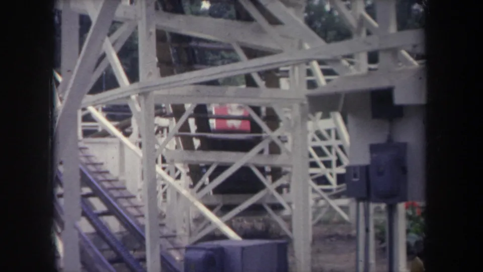 1968 Michigan home-movie view of a red roller coaster train moving through white wooden supports, with riders visible at a distance.