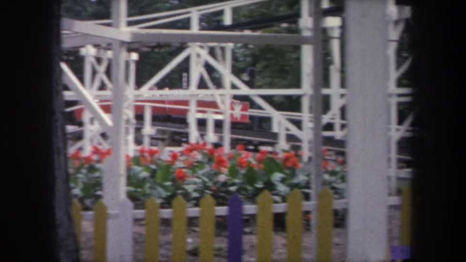 1968 Michigan amusement park frame showing red flowers, a low picket fence, white coaster supports, and a red train in the background.