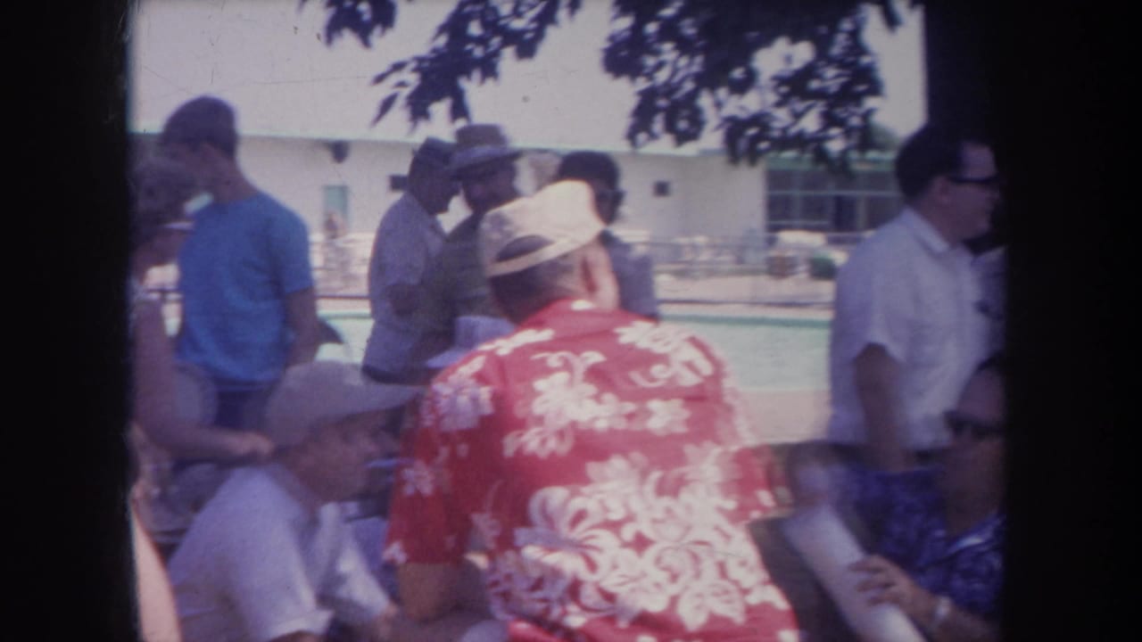 1968 Michigan home-movie frame showing men socializing beside a swimming pool, including a red floral shirt, hats, and shaded picnic seating.