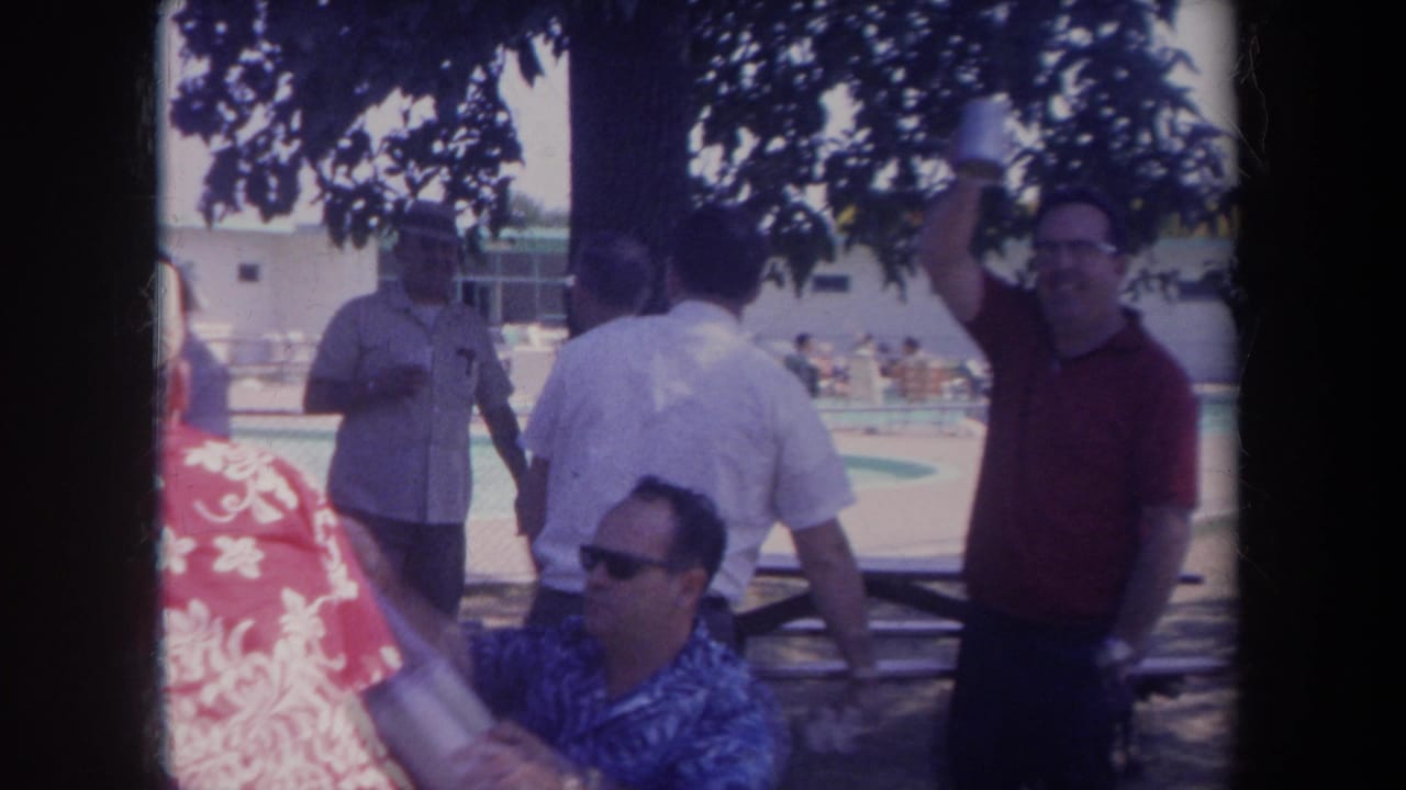 Men stand and sit near picnic tables beside a pool, with a man in a red shirt at right.
