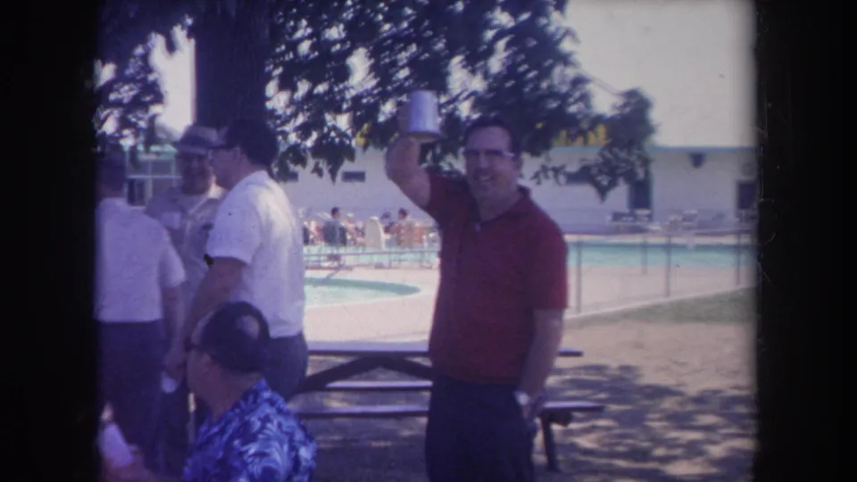 1968 Michigan home-movie frame of a man raising a metal cup near shaded poolside seating while others socialize nearby.