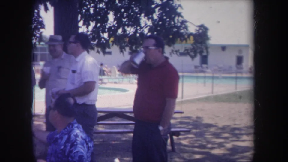 1968 Michigan poolside home-movie frame showing a man in a red shirt drinking from a cup while other men stand near picnic tables.