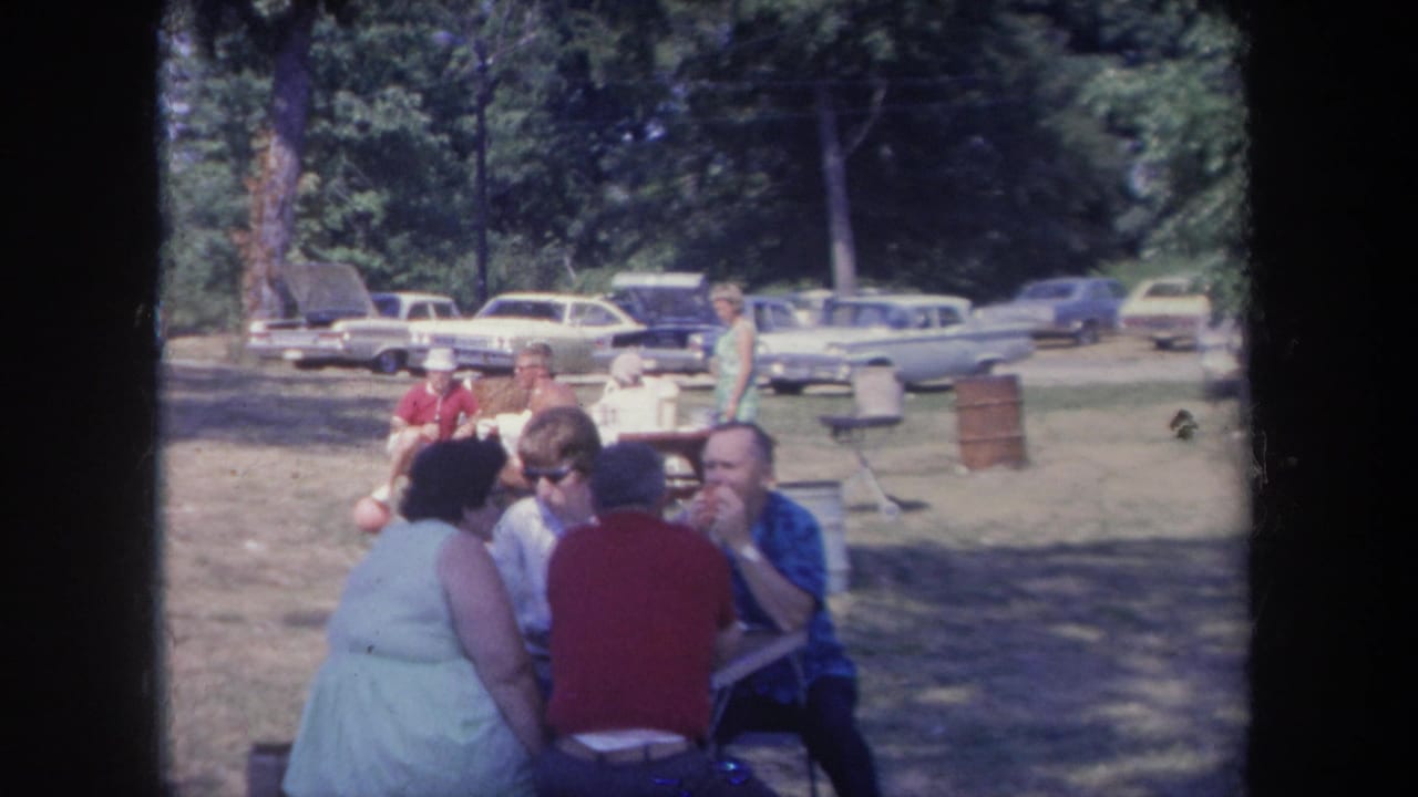 1968 Michigan home-movie frame showing adults eating at an outdoor picnic table with 1960s cars parked under trees in the background.