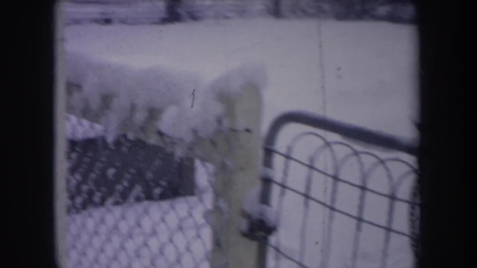 1968 Michigan home-movie frame showing a snowy backyard, chain-link fence, house siding and a dark dog partly visible near the snow.
