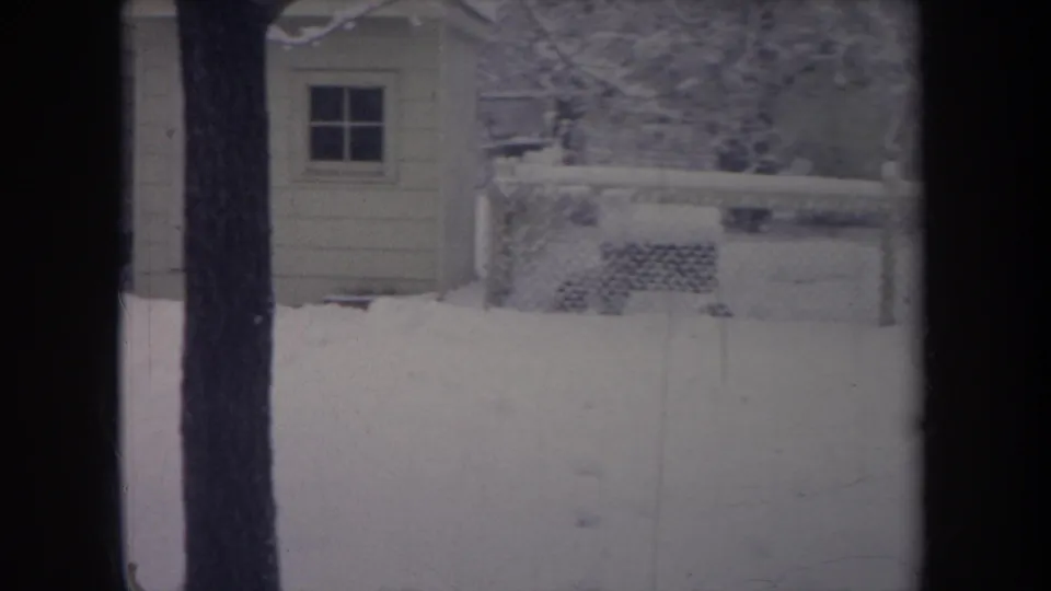 1968 Michigan 8mm frame of a winter backyard with a small shed, chain-link fence, tree trunk, and accumulated snow.