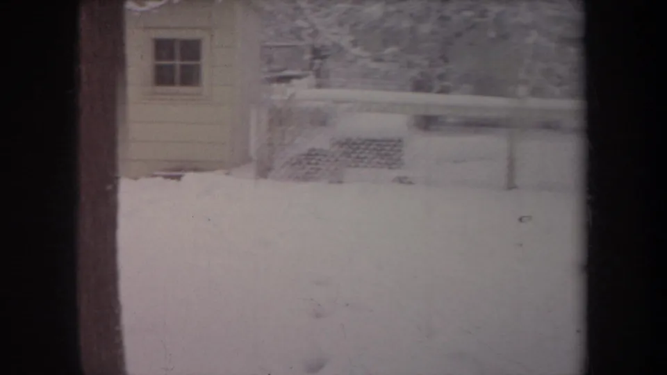 1968 Michigan home-movie frame with a snowy fenced yard, small shed, and a small dog partly visible low in the snow.