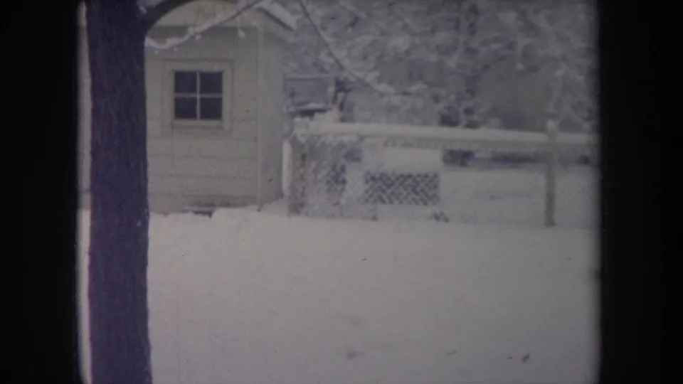 1968 Michigan home-movie frame of a snow-covered backyard viewed through a window, with a pale shed, fence, and bare trees.