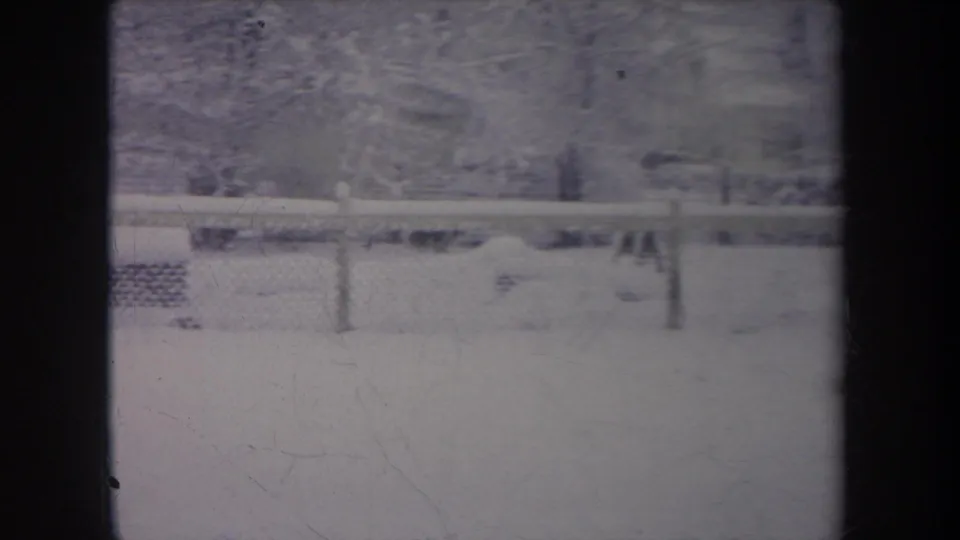 1968 Michigan backyard in deep snow, filmed through a dark window frame with fence posts and trees visible beyond.