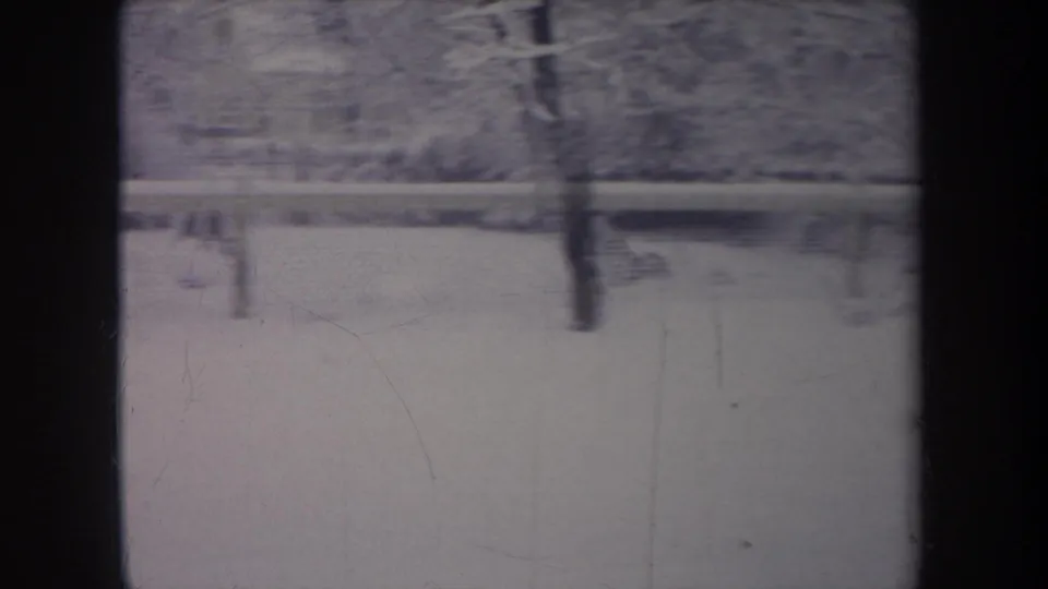 1968 Michigan home-movie frame of a quiet snow-covered backyard through a window, with fence posts, trees, and soft residential background detail.