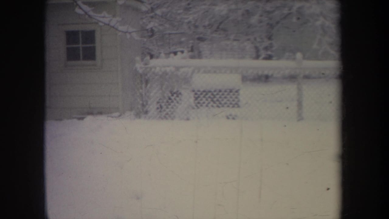 1968 Michigan home-movie frame showing a snow-covered residential yard with chain-link fence, trees, and a small structure at the left edge.