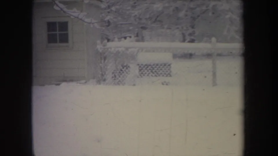 1968 Michigan home-movie frame showing a snow-covered residential yard with chain-link fence, trees, and a small structure at the left edge.
