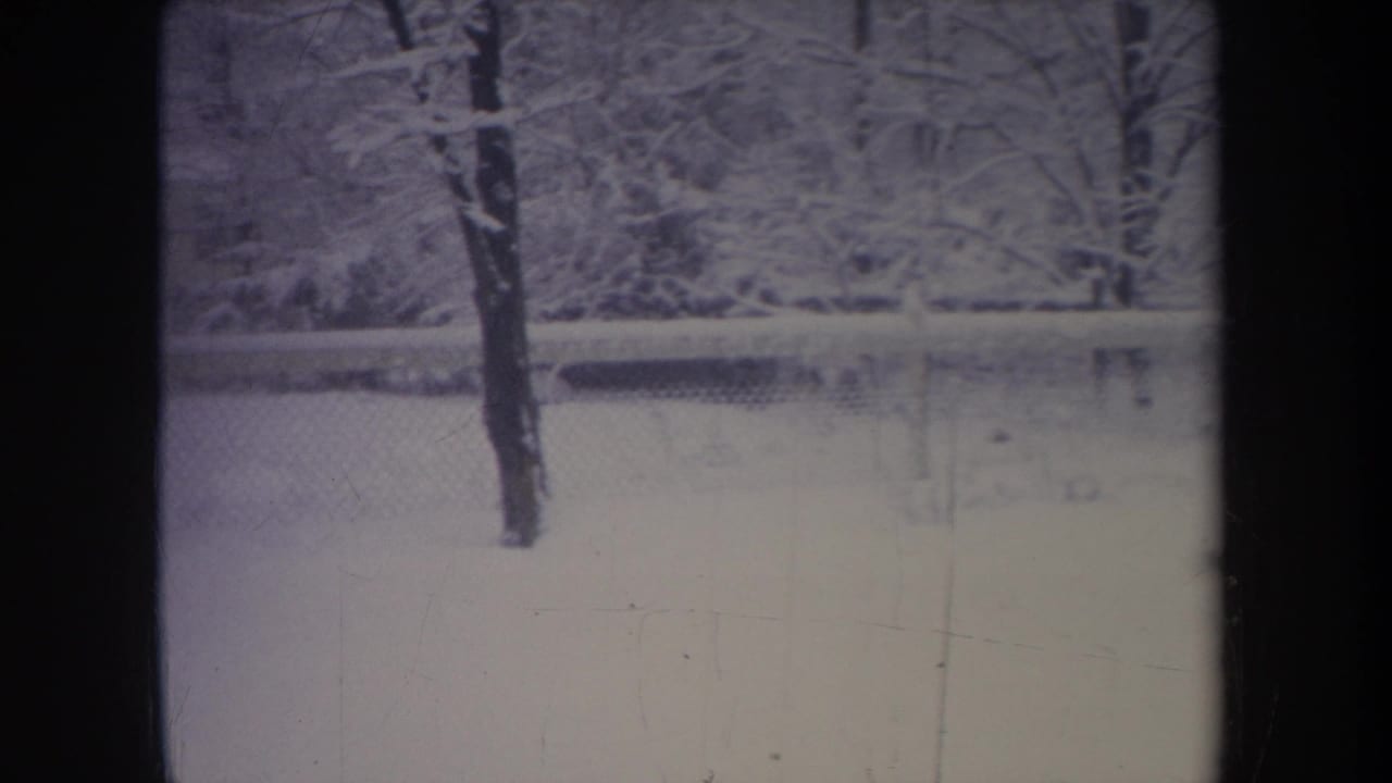1968 Michigan 8mm frame showing a snow-covered residential yard through dark window mullions, with bare trees and a chain-link fence.