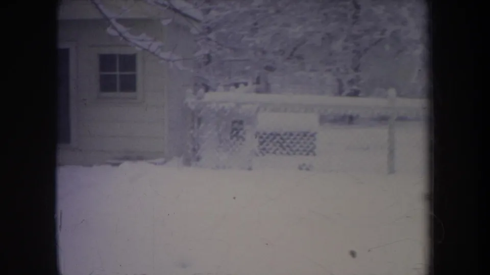 1968 Michigan home-movie frame showing a small house wall and window beside a snow-covered fenced backyard.