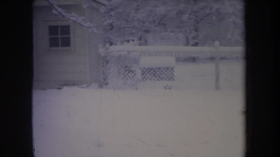1968 Michigan 8mm frame showing a snow-covered yard, small outbuilding, fence, and bare branches in soft focus.