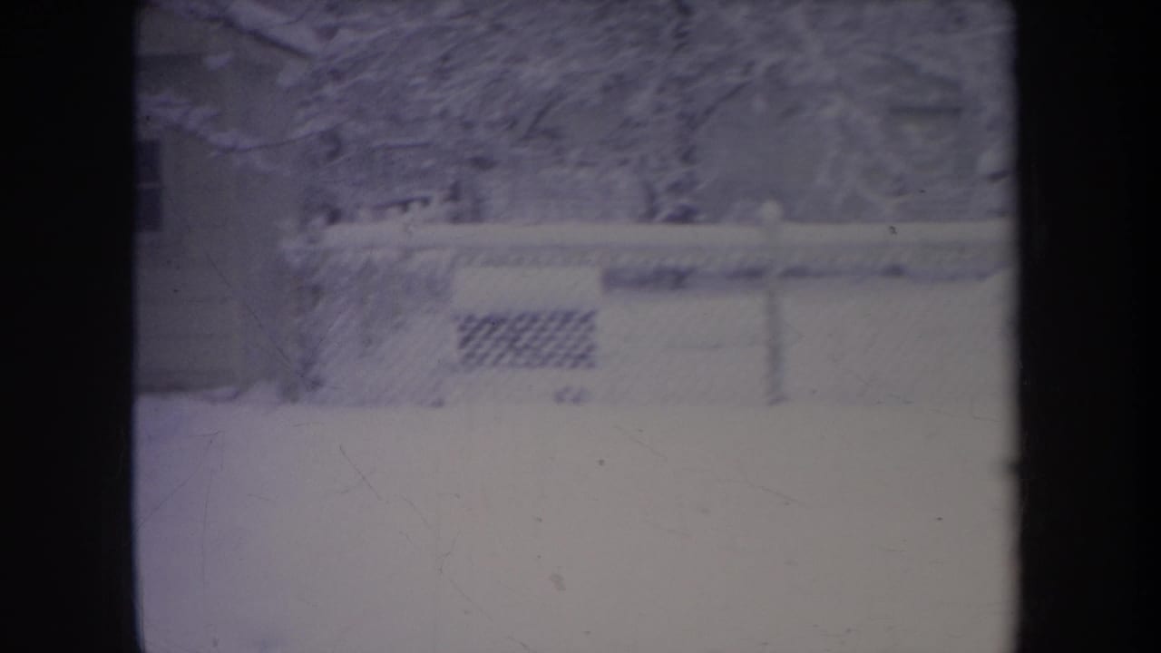 Snowy fenced backyard with bare trees and a house exterior behind dark film borders.