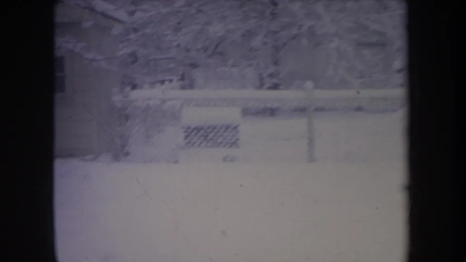 1968 Michigan home-movie frame showing a snow-covered residential yard, chain-link fence, trees, and a house wall in soft 8mm color.