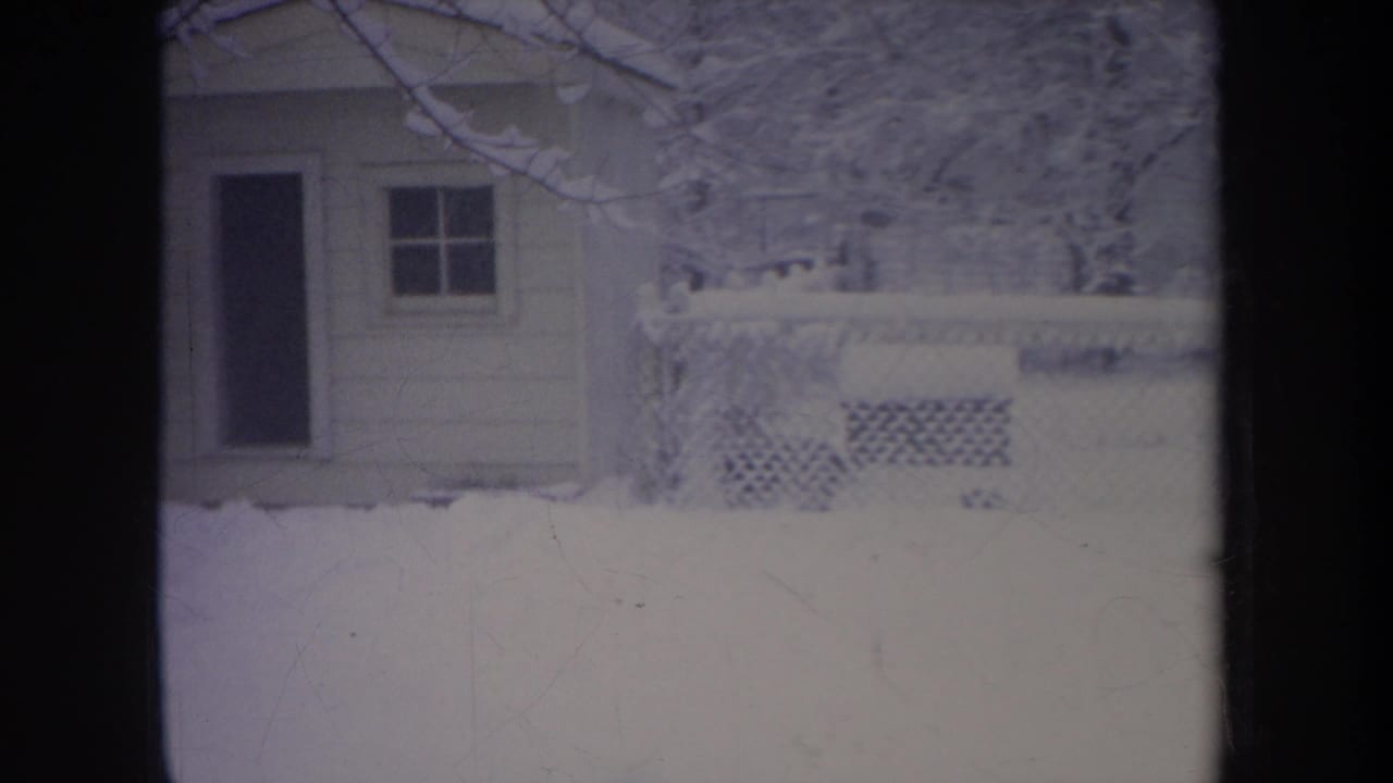 Small house in deep snow with a door, window, fence, and bare branches.