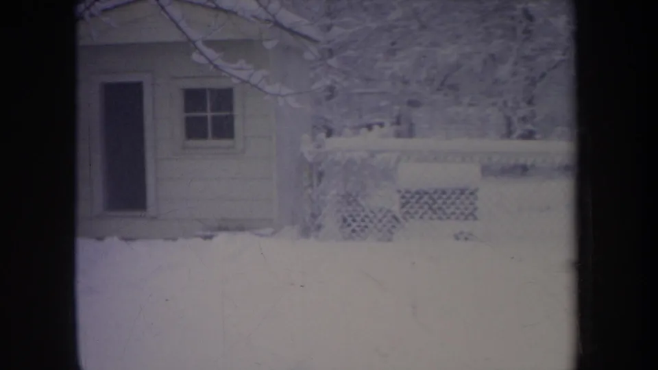1968 Michigan winter frame with a small house partly framed by snow, chain-link fencing, bare branches, and a dark door.