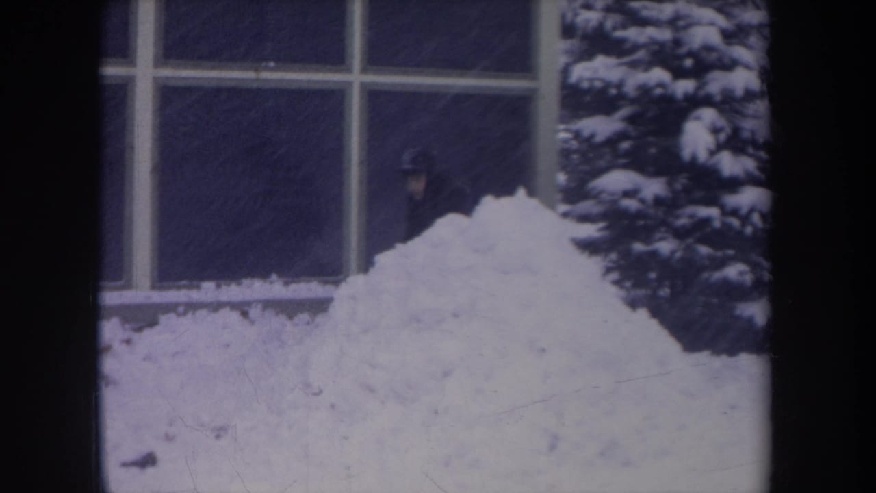 1968 Michigan 8mm frame with a bundled person near a large window, snow pile, and snow-covered evergreen shrub.