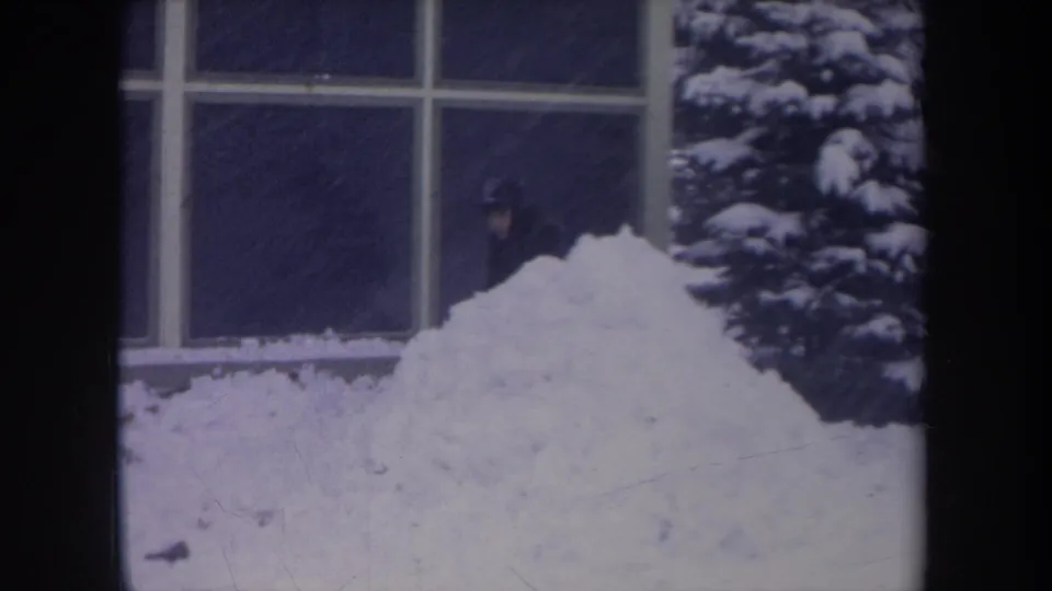 1968 Michigan 8mm frame with a bundled person near a large window, snow pile, and snow-covered evergreen shrub.