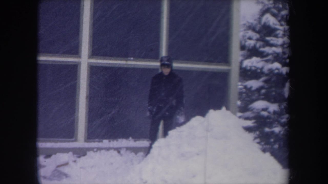 1968 Michigan winter frame showing a bundled person near large house windows, deep snow, and a snow-covered evergreen.