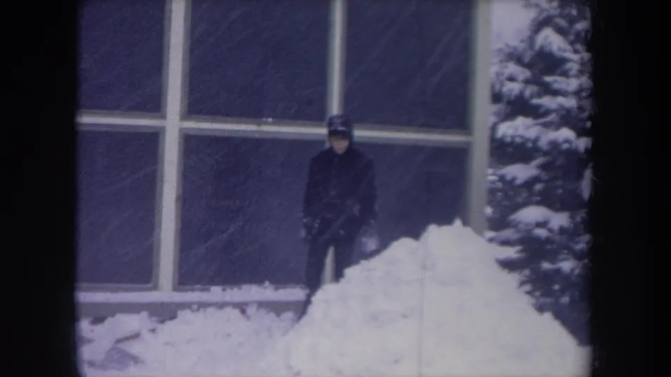 1968 Michigan winter frame showing a bundled person near large house windows, deep snow, and a snow-covered evergreen.