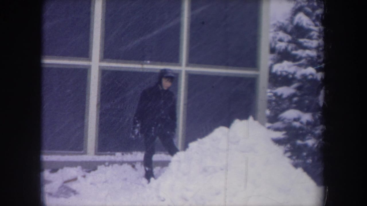 Young man in dark winter clothes stands beside a deep snowbank in front of large grid windows.