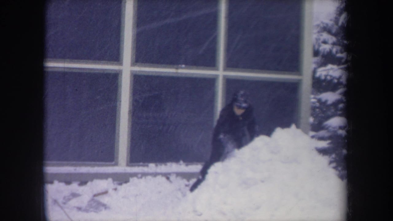 Dark-clad figure bends near a snowbank beside a snow-covered evergreen and large windows.