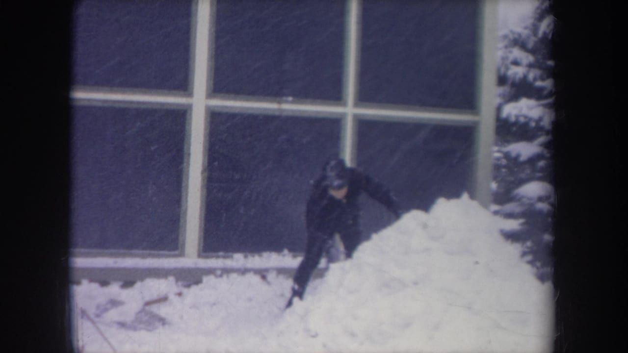 A young man in dark winter clothing reaches down toward deep snow below large windows in the 1968 Michigan scene.
