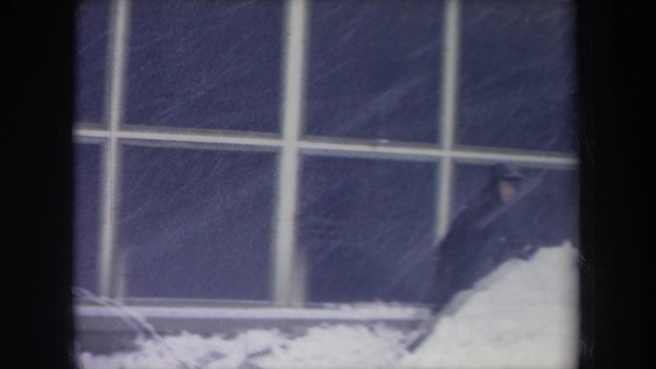 The figure is partly cropped at frame right while working near a snowbank beneath large grid windows in the Michigan winter clip.