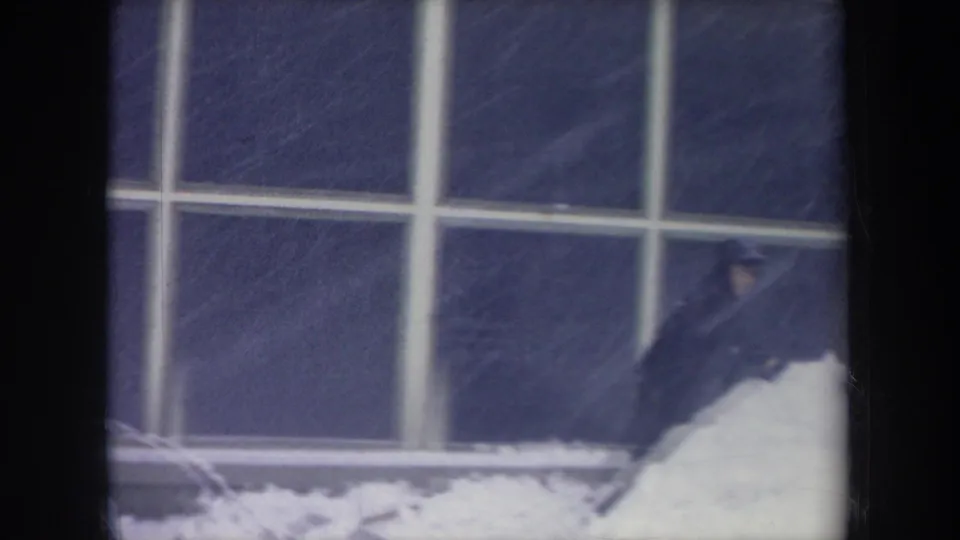 The figure is partly cropped at frame right while working near a snowbank beneath large grid windows in the Michigan winter clip.
