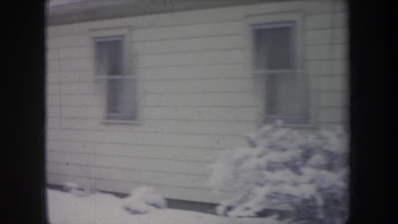 1968 Michigan 8mm frame of a snow-covered residential exterior with windows, siding, and shrubs.