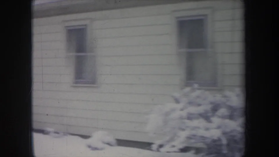 1968 Michigan 8mm frame of a snow-covered residential exterior with windows, siding, and shrubs.