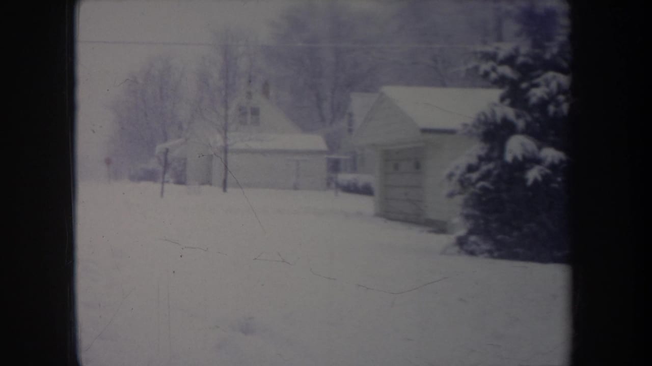 Snowy yard seen through a window with a garage, houses, and an evergreen tree.