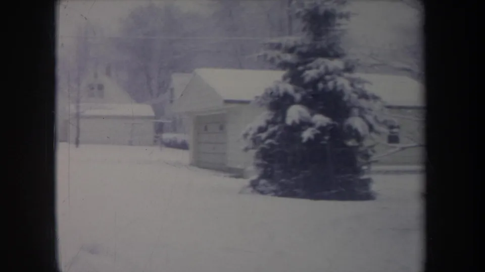 1968 Michigan winter frame with snow-covered houses, a detached garage, bare trees, and a large evergreen seen from indoors.