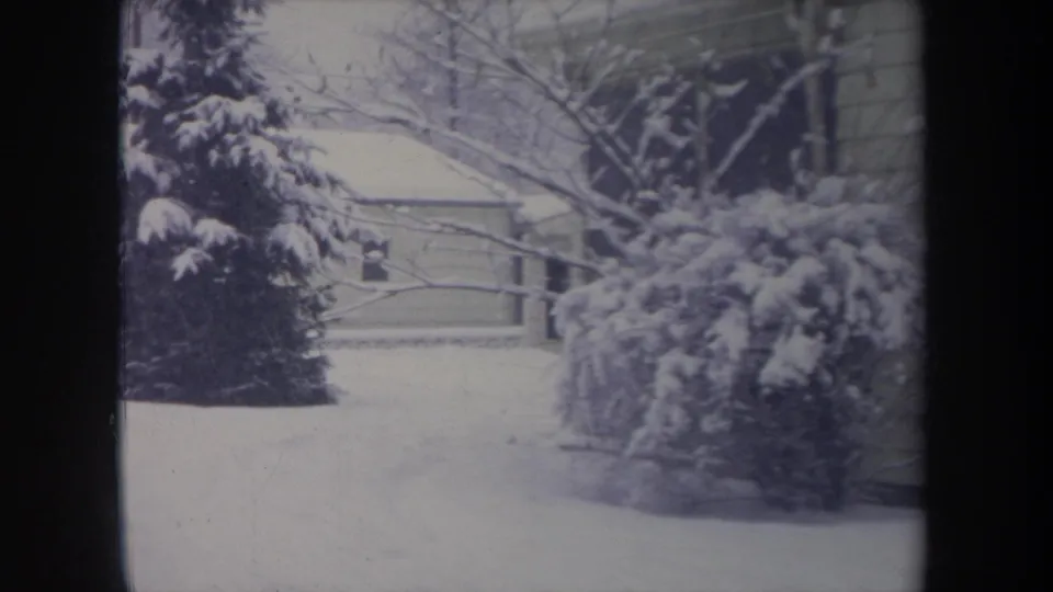 1968 Michigan suburban winter frame showing snow-laden shrubs, bare branches, nearby houses, overhead lines, and a pale snow-covered lawn.