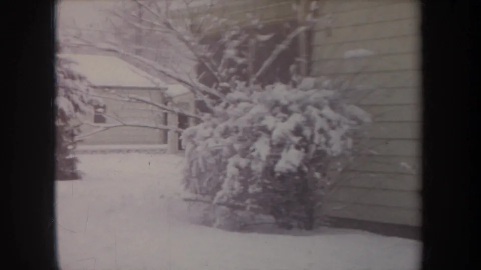 1968 Michigan home-movie frame showing snow-laden shrubs, bare branches, a snowy yard, and the side of a pale residential house.