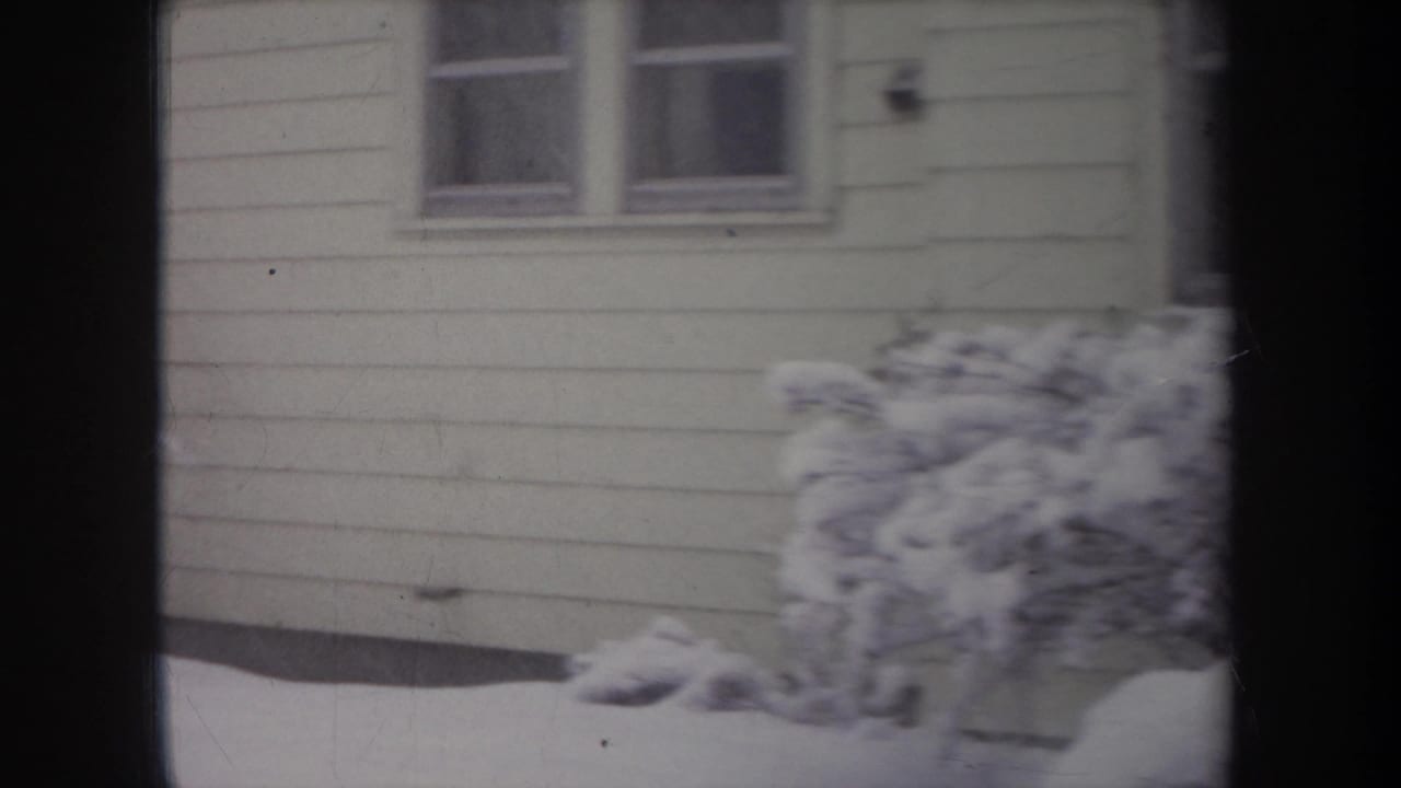 1968 Michigan home-movie frame showing pale siding, two house windows, deep snow, and shrubs partly buried by winter accumulation.