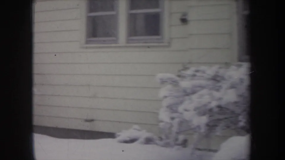 1968 Michigan home-movie frame showing pale siding, two house windows, deep snow, and shrubs partly buried by winter accumulation.