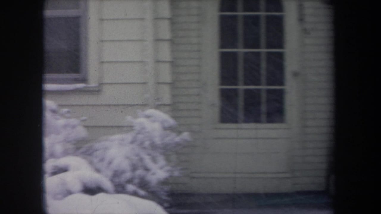 Snowy house exterior seen through dark window framing, with a door, window, and snow-covered shrubs.
