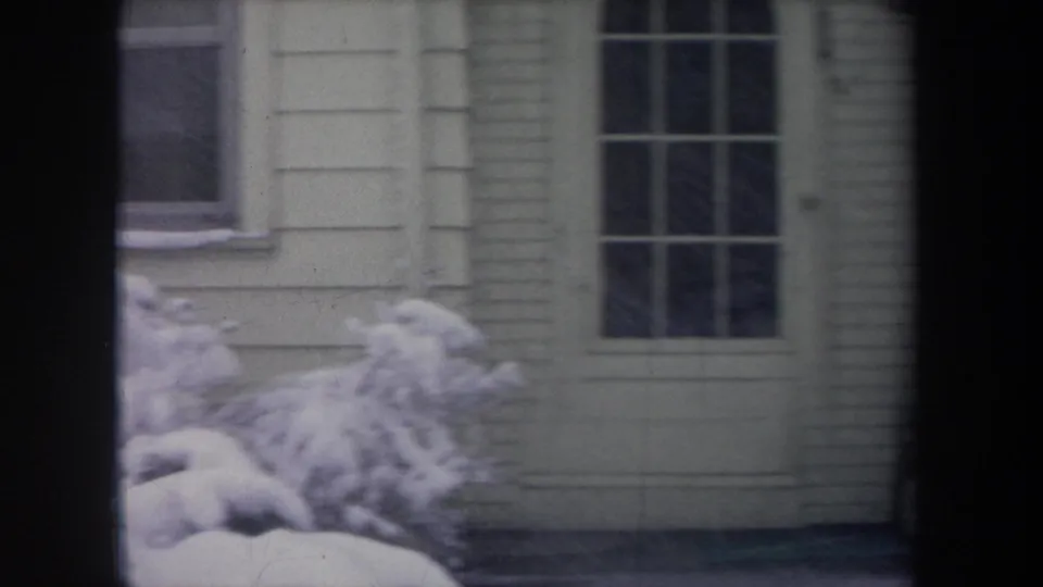 1968 Michigan 8mm frame showing a white-sided house exterior through dark window framing, with snow on shrubs and the yard.