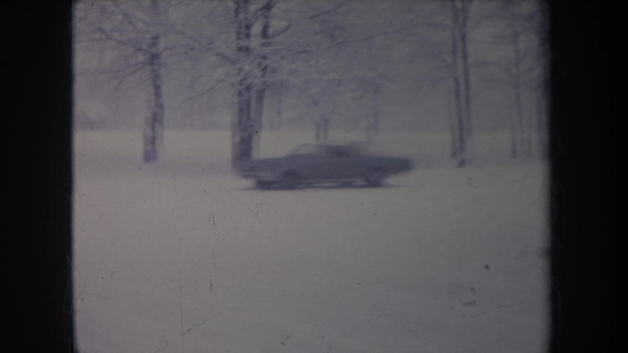 A dark car moves through a snow-covered tree-lined area in 1968 Michigan home-movie footage.