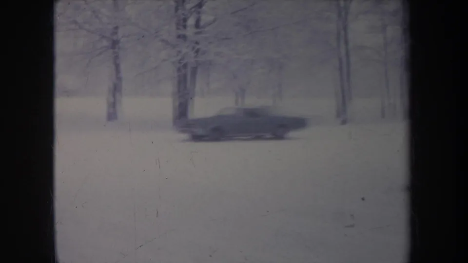 A dark car moves through a snow-covered tree-lined area in 1968 Michigan home-movie footage.