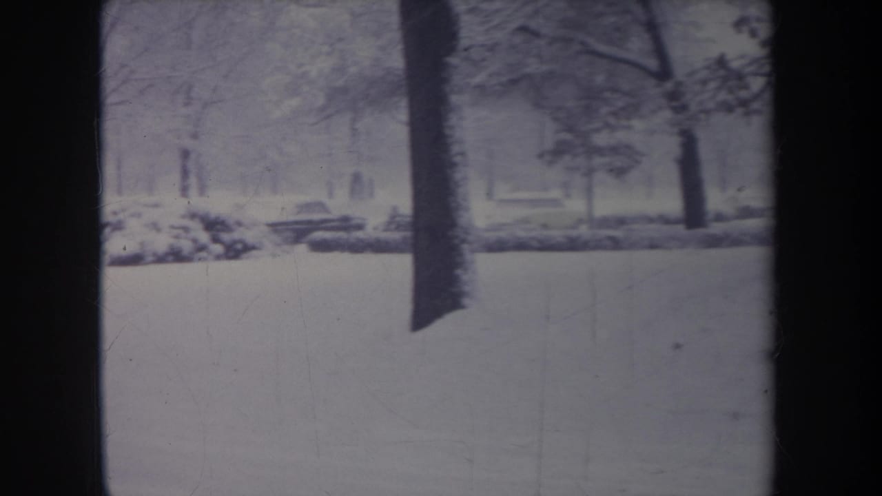 1968 Michigan 8mm frame with a foreground tree trunk dividing a snowy yard as the older car passes in back.