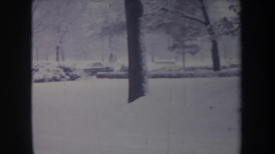 1968 Michigan 8mm frame with a foreground tree trunk dividing a snowy yard as the older car passes in back.