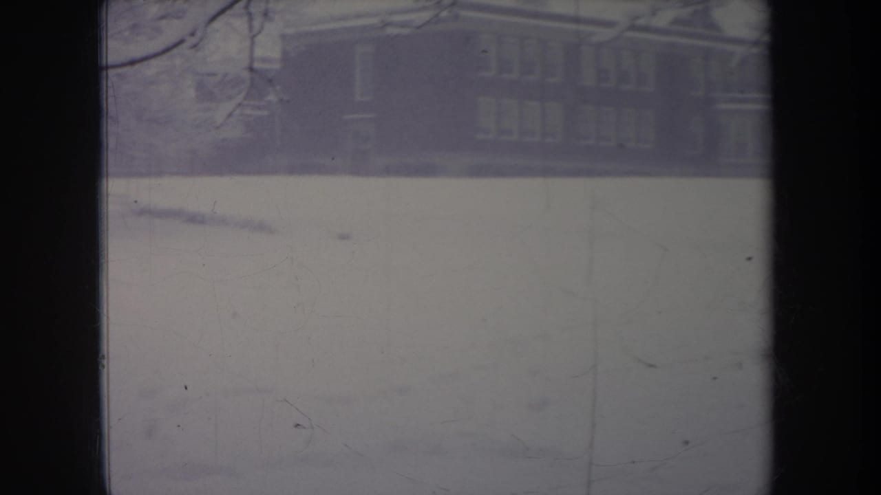 1968 Michigan 8mm frame of a large windowed house behind a snow-covered yard and bare winter branches.