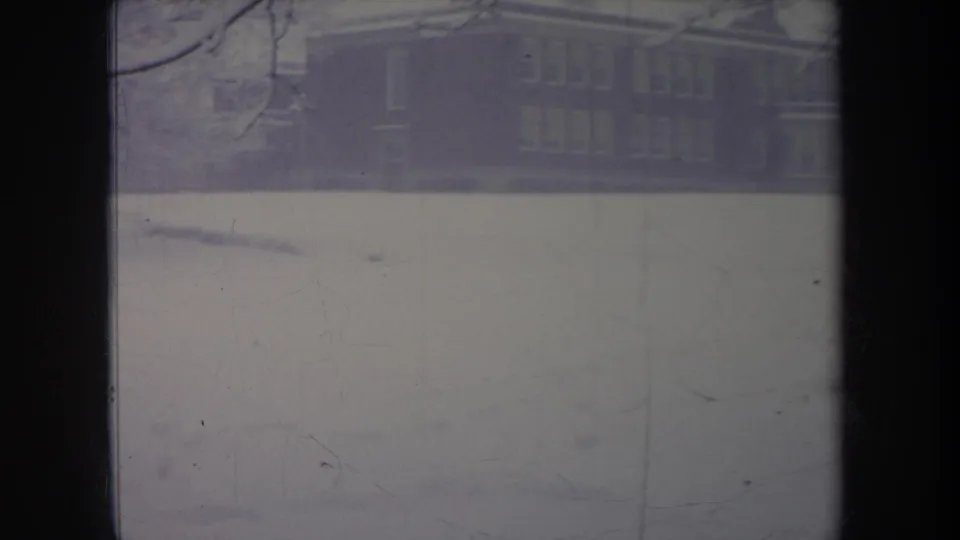 1968 Michigan 8mm frame of a large windowed house behind a snow-covered yard and bare winter branches.
