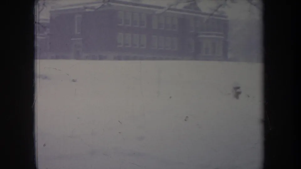 1968 Michigan 8mm frame showing a snowy open area before a brick building, with a small dog moving across the foreground.