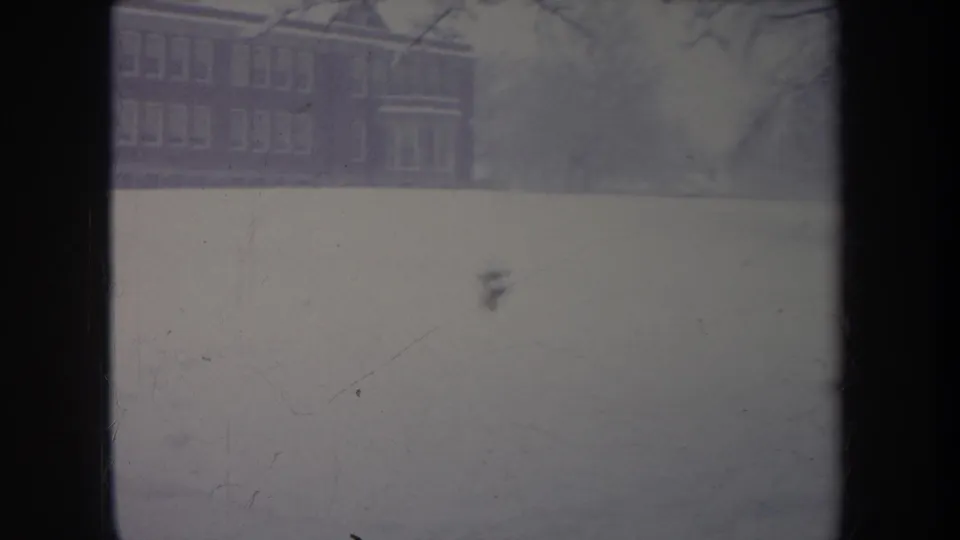 1968 Michigan winter frame with a distant brick building and a small dog moving across the snow-covered grounds.